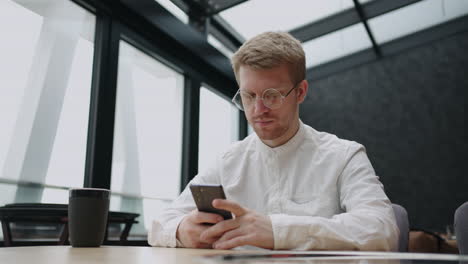 A-young-man-with-glasses-with-a-mobile-phone-in-his-hands-looks-at-the-screen-and-prints-a-message.-Hipster-in-round-glasses-writes-a-message-on-his-phone